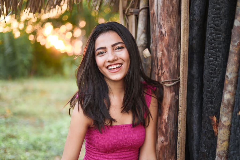 Yolane is wearing a pink dress. She is leaning against a wooden building.